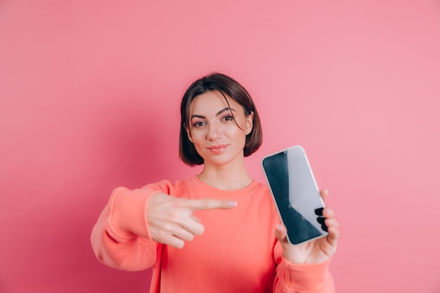 ¡Mira este celular! La mujer feliz y complacida señala con el dedo índice en la pantalla en blanco, muestra un dispositivo moderno, emociones felices y sorprendidas.