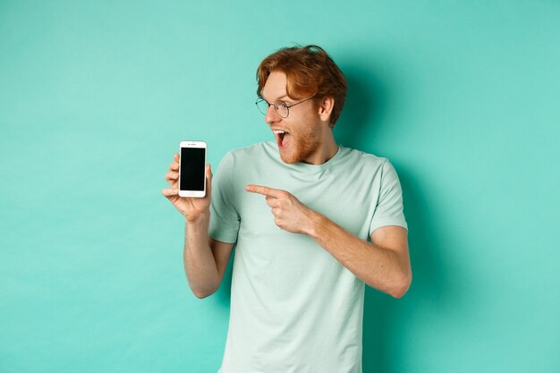 Mira esto. Chico guapo pelirrojo con gafas apuntando con el dedo a la pantalla del teléfono inteligente en blanco, mostrando promoción en línea, de pie asombrado sobre fondo turquesa.