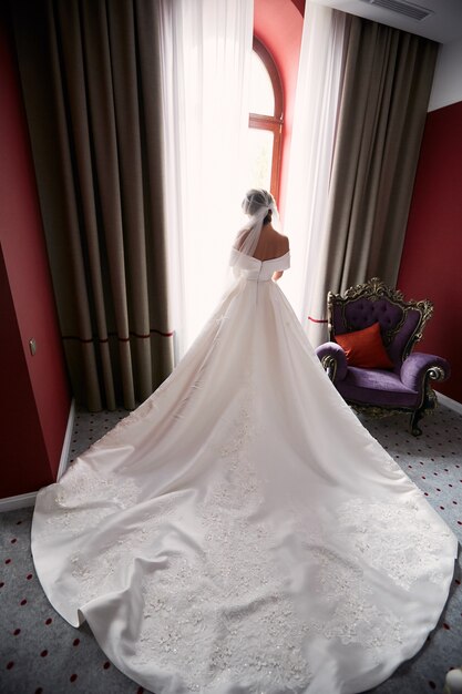 Mira por detrás a la novia con vestido largo parado frente a la ventana.