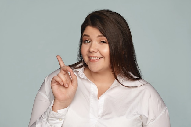 Foto gratuita mira esto. juguetona hermosa joven con cabello negro suelto y kilos de más guiñando un ojo y sonriendo, apuntando con el dedo índice a la pared en blanco con espacio de copia para su contenido