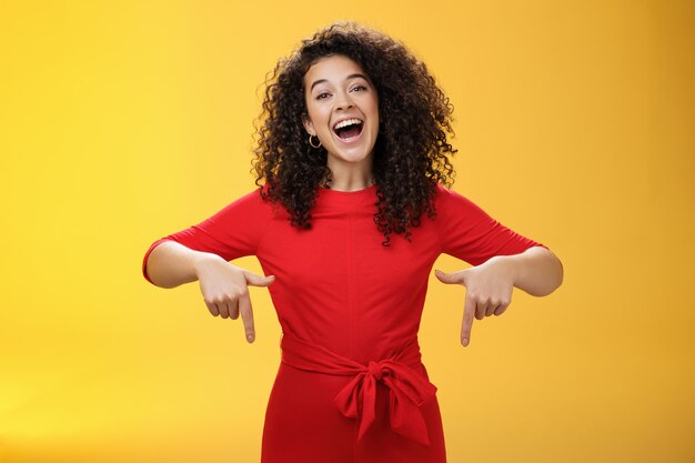 Mira lo que tengo aquí. Carismática despreocupada feliz encantadora mujer con cabello rizado en vestido rojo riendo con una amplia sonrisa apuntando hacia abajo como mostrando un impresionante espacio de copia a los clientes sobre la pared amarilla.