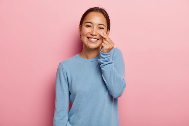 Mira mi mejilla. Mujer asiática de aspecto agradable toca la piel de la cara, demuestra su frescura y suavidad, no usa maquillaje, cabello oscuro peinado, vestida con una sudadera azul informal, aislada en rosa