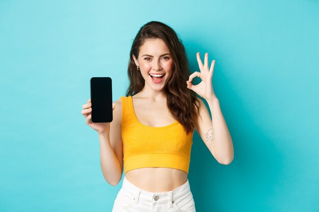 Mira esto. Mujer guapa con sonrisa descarada, guiñando un ojo y mostrando un signo de bien con la pantalla vacía del teléfono inteligente, recomendando la aplicación, de pie sobre fondo azul.