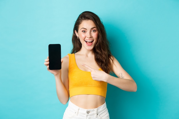 Mira esto. Mujer joven sonriente apuntando a la pantalla vacía del teléfono inteligente, recomendando promoción en línea, de pie contra el fondo azul.