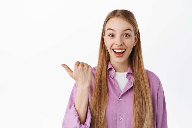 Mira esto Sonriente mujer feliz con cabello largo y rubio apuntando con el pulgar hacia la izquierda anunciando descuentos especiales que muestran ofertas promocionales de pie contra fondo blanco