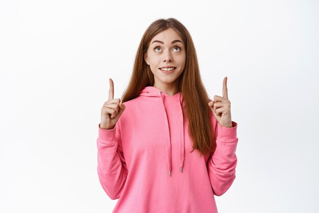 Mirada de adolescente con cara esperanzada en la parte superior sonriendo y señalando con el dedo de pie en ropa informal contra el fondo blanco.