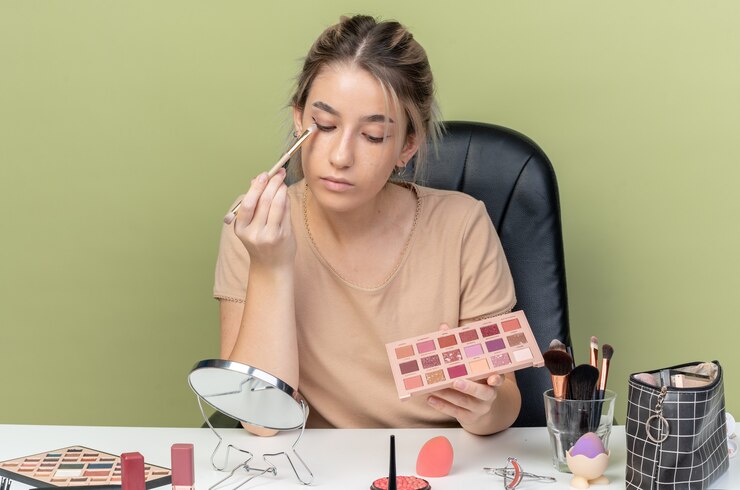 Foto gratuito mirando al espejo hermosa joven sentada en el escritorio con herramientas de maquillaje aplicando sombra de ojos con pincel de maquillaje aislado en la pared verde oliva