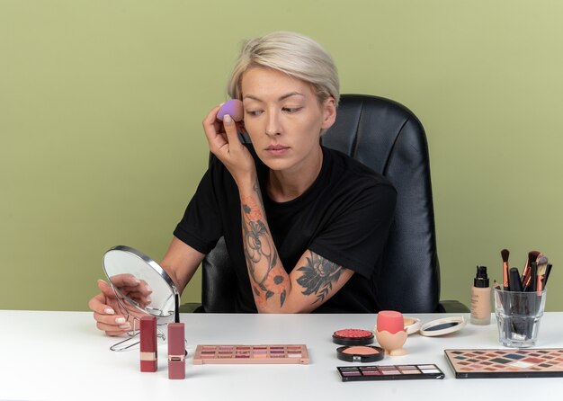 Mirando al espejo joven hermosa se sienta a la mesa con herramientas de maquillaje aplicando crema tonificante con esponja aislada en la pared verde oliva