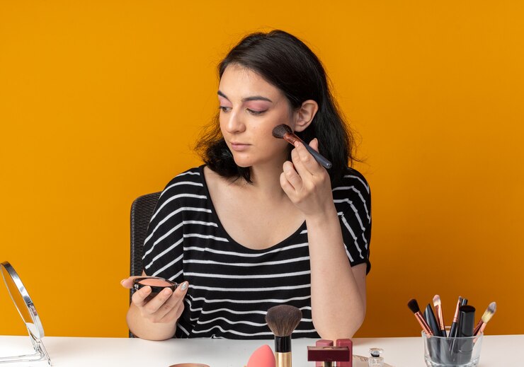Foto gratuito mirando al espejo joven hermosa se sienta a la mesa con herramientas de maquillaje aplicando rubor en polvo con pincel aislado en pared naranja