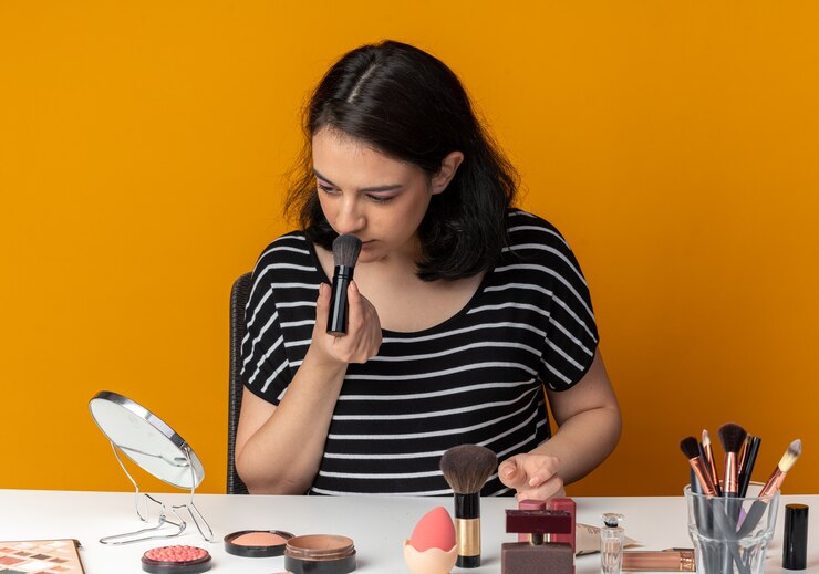 Foto gratuito mirando al espejo joven hermosa se sienta a la mesa con herramientas de maquillaje aplicando rubor en polvo con pincel aislado en pared naranja