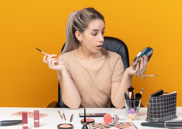 Mirando al espejo joven hermosa se sienta a la mesa con herramientas de maquillaje aplicando sombra de ojos con pincel de maquillaje aislado en la pared naranja