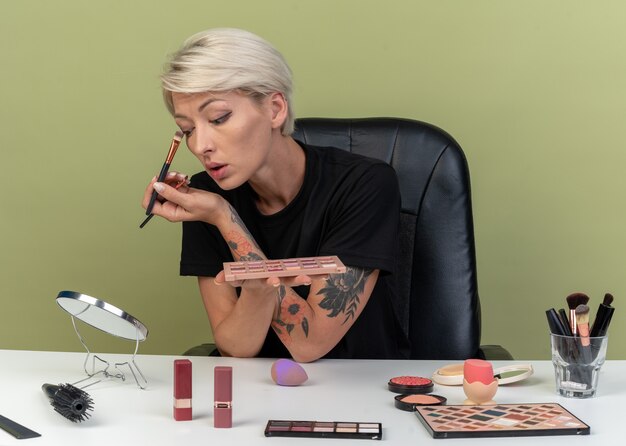 Mirando al espejo joven hermosa se sienta a la mesa con herramientas de maquillaje aplicando sombra de ojos con pincel de maquillaje aislado en la pared verde oliva