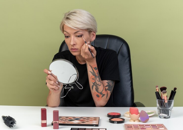 Foto gratuito mirando al espejo joven hermosa se sienta a la mesa con herramientas de maquillaje aplicando sombra de ojos con pincel de maquillaje aislado en la pared verde oliva