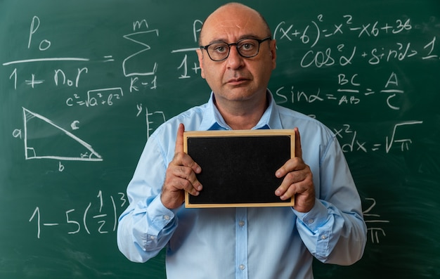 Mirando al frente profesor de mediana edad con gafas de pie delante de la pizarra sosteniendo una mini pizarra