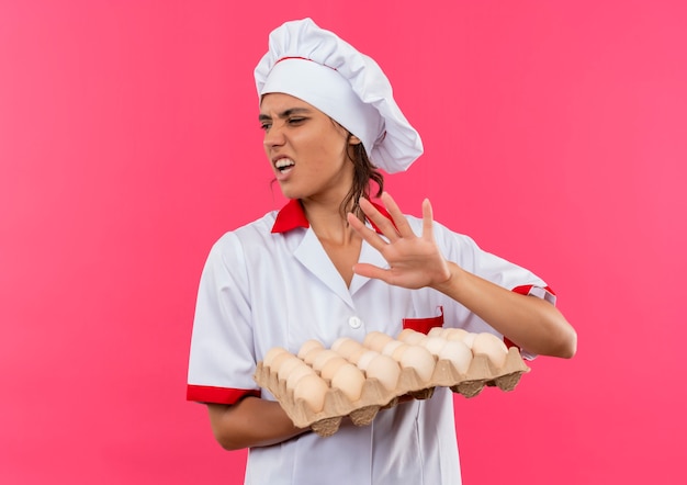 Foto gratuita mirando al lado aprensivo joven cocinera vistiendo uniforme de chef sosteniendo un lote de huevos en la pared rosa aislada con espacio de copia