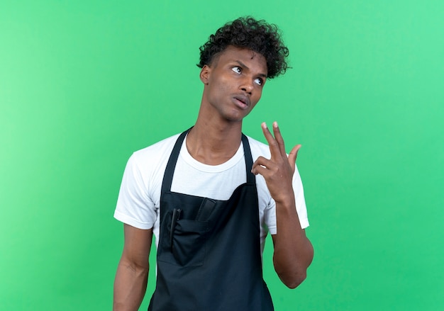 Mirando al lado cansado joven peluquero masculino afroamericano vistiendo uniforme mostrando gesto de pistola aislado sobre fondo verde