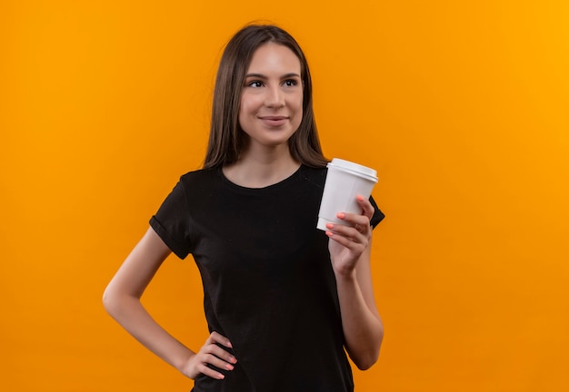 Mirando al lado complacido joven caucásica vestida con camiseta negra sosteniendo una taza de café puso su mano en la cadera en la pared naranja aislada