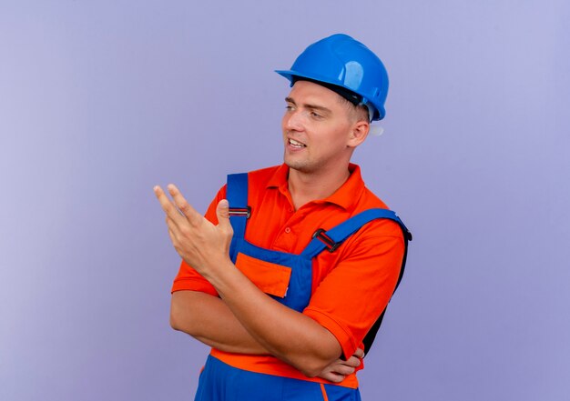 Mirando al lado complacido joven constructor masculino vistiendo uniformes y puntos de casco de seguridad con la mano al lado