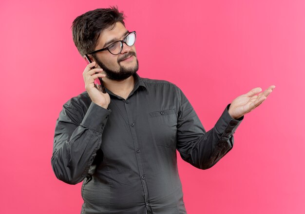 Mirando al lado complacido joven empresario con gafas habla por teléfono y señala con la mano al lado aislado en la pared rosa