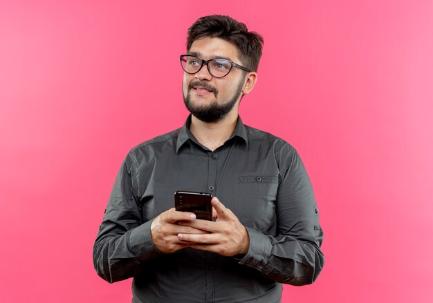 Mirando al lado complacido joven empresario con gafas sosteniendo teléfono aislado sobre fondo de color rosa