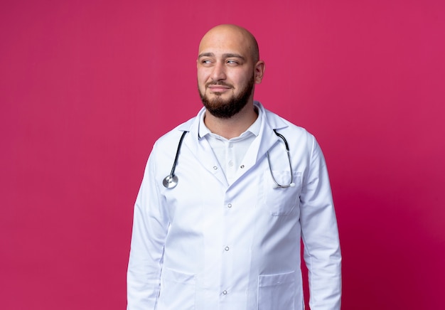 Foto gratuita mirando al lado complacido joven médico varón calvo vistiendo bata médica y estetoscopio aislado sobre fondo rosa con espacio de copia