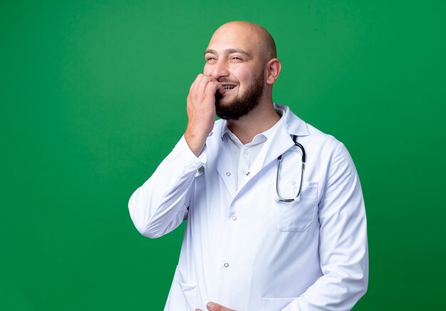 Mirando al lado complacido joven médico vistiendo bata médica y estetoscopio muerde las uñas aisladas sobre fondo verde