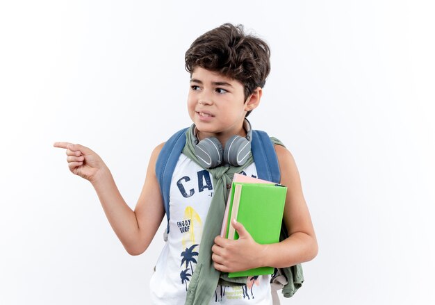 Mirando al lado complacido pequeño colegial con mochila y auriculares sosteniendo libro y puntos en el lado aislado sobre fondo blanco con espacio de copia