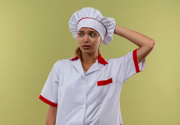 Mirando al lado confundido joven cocinera vistiendo uniforme de chef poniendo la mano en la cabeza en la pared verde aislada con espacio de copia