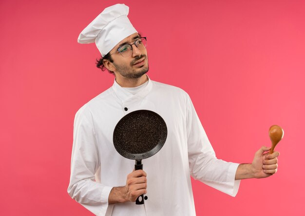 Mirando al lado confundido joven cocinero con uniforme de chef y gafas sosteniendo una sartén y una cuchara