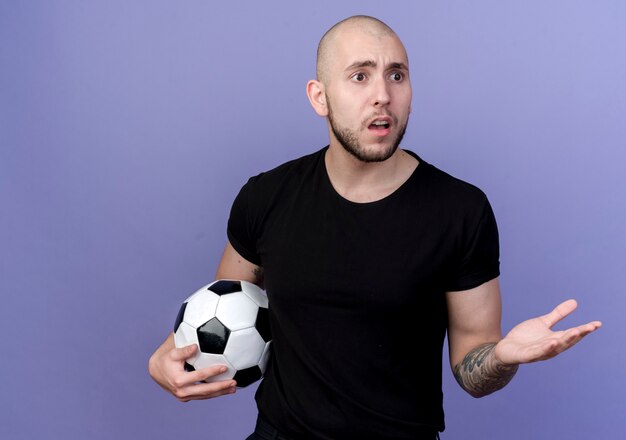 Mirando al lado confundido joven deportivo sosteniendo la bola y los puntos con la mano al lado aislado sobre fondo púrpura