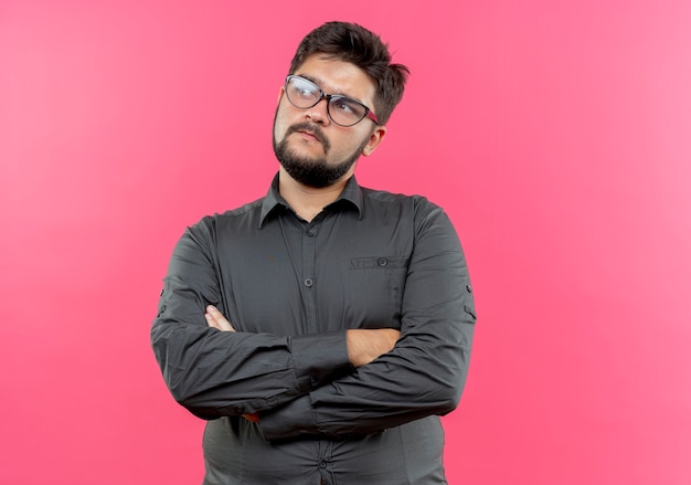 Mirando al lado confundido joven empresario con gafas cruzando las manos aisladas en la pared rosa