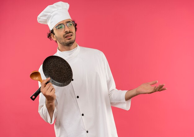 Mirando al lado descontento joven cocinero vistiendo uniforme de chef y gafas sosteniendo una sartén con cuchara y mano extendida