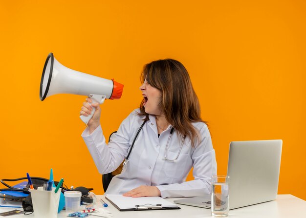 Mirando al lado de la doctora de mediana edad con bata médica con estetoscopio sentado en el escritorio, trabajo en una computadora portátil con herramientas médicas, habla por un altavoz en una pared naranja aislada con espacio de copia