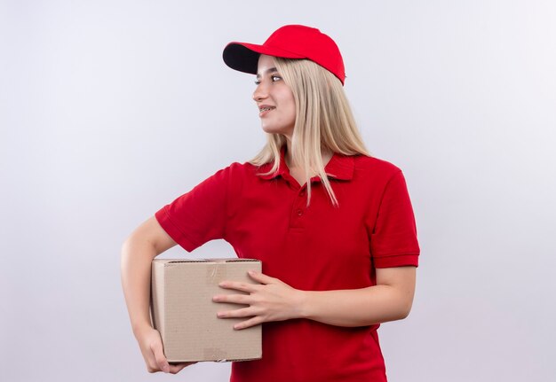 Mirando al lado de la entrega joven vistiendo camiseta roja y gorra con caja en pared blanca aislada