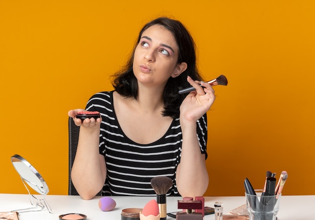 Mirando al lado hermosa joven se sienta a la mesa con herramientas de maquillaje sosteniendo rubor en polvo con pincel aislado en la pared naranja