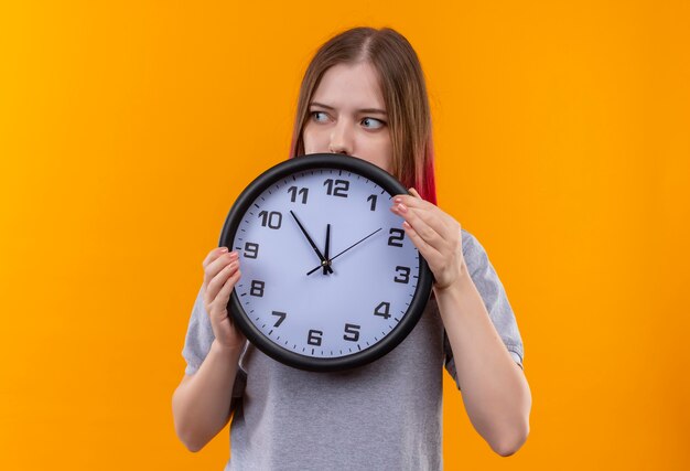 Mirando al lado hermosa joven vestida con camiseta gris con reloj de pared sobre fondo amarillo aislado