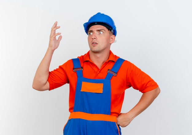 Mirando al lado impresionado joven constructor vistiendo uniforme y casco de seguridad levantando la mano poniendo en la cadera otra mano