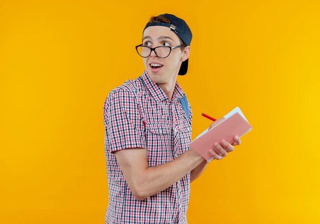 Mirando al lado impresionado joven estudiante chico con gafas y gorra escribiendo algo en el cuaderno