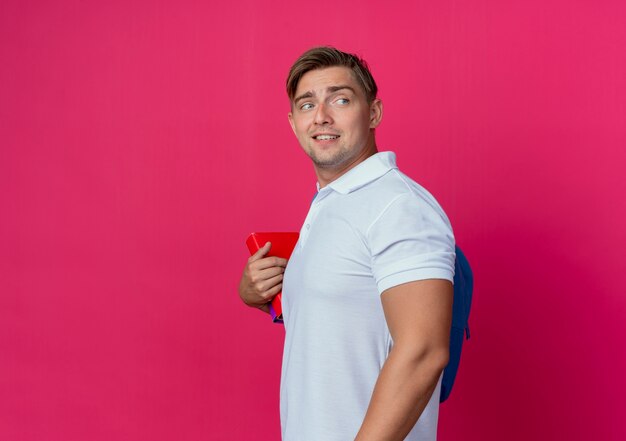 Mirando al lado joven apuesto estudiante masculino con bolsa trasera sosteniendo libros aislados en la pared rosa