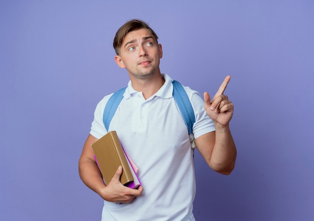 Mirando al lado joven apuesto estudiante masculino con mochila sosteniendo libros y puntos en el lado aislado en la pared azul con espacio de copia