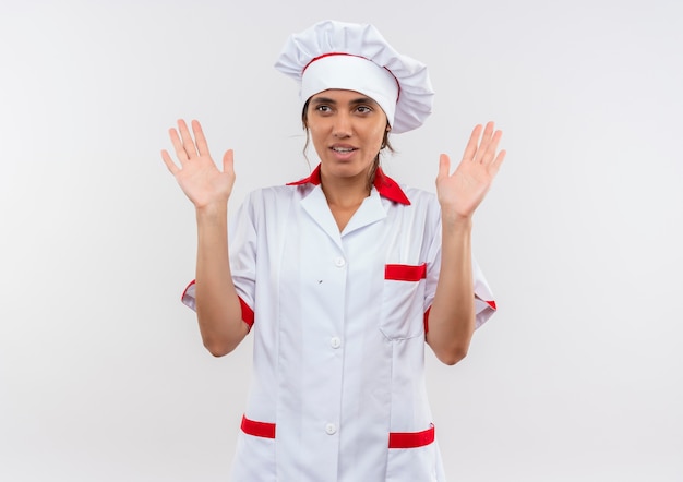 Mirando al lado joven cocinera vistiendo uniforme de chef que muestra el tamaño en la pared blanca aislada con espacio de copia