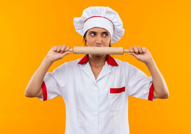 Mirando al lado joven cocinera vistiendo uniforme de chef sosteniendo y muerde el rodillo en la pared amarilla aislada con espacio de copia