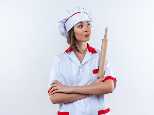 Mirando al lado joven cocinera vistiendo uniforme de chef sosteniendo un rodillo cruzando las manos aisladas en la pared blanca