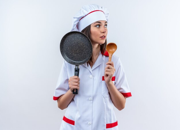 Mirando al lado joven cocinera vistiendo uniforme de chef sosteniendo una sartén con cuchara aislado en la pared blanca