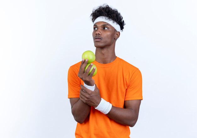 Mirando al lado joven deportivo afroamericano vistiendo diadema y muñequera sosteniendo manzanas aisladas sobre fondo blanco.