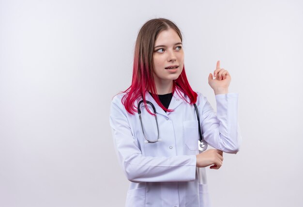 Mirando al lado joven médico chica con estetoscopio bata médica apunta hacia arriba sobre fondo blanco aislado