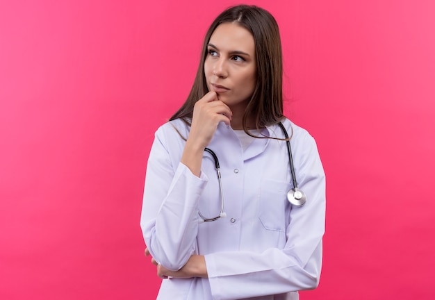 Foto gratuita mirando al lado joven médico chica con estetoscopio bata médica puso su mano en la barbilla sobre fondo rosa aislado