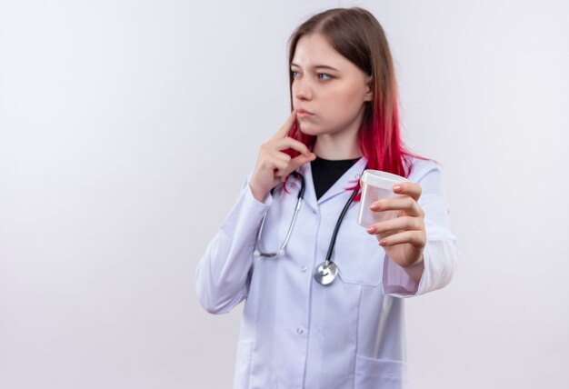 Mirando al lado joven médico chica con estetoscopio bata médica sosteniendo lata vacía a la cámara sobre fondo blanco aislado