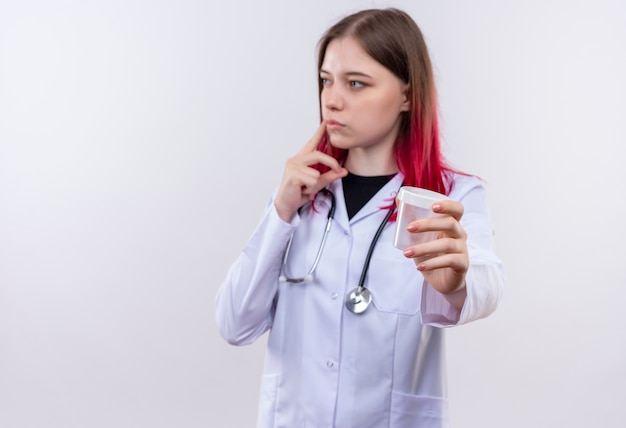 Mirando al lado joven médico chica con estetoscopio bata médica sosteniendo lata vacía a la cámara sobre fondo blanco aislado