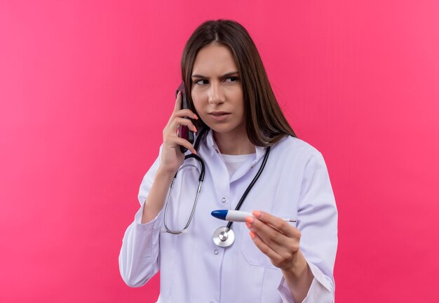 Mirando al lado joven médico chica con estetoscopio bata médica sosteniendo termómetro habla por teléfono sobre fondo rosa aislado